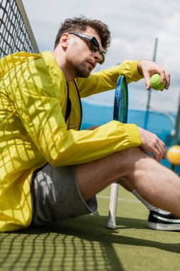 Güneş gözlüklü erkek oyuncu tenis ağının yanında oturuyor, elinde top ve tenis raketi, spor ve stil var.
