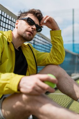 Güneş gözlüklü yakışıklı erkek oyuncu tenis ağının yanında oturuyor, topu tutuyor, spor ve tarz sahibi.