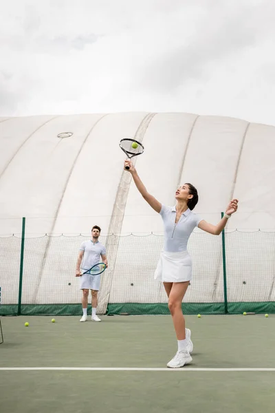 athletic woman in active wear hitting ball with racket, man standing on court, motivation and sport