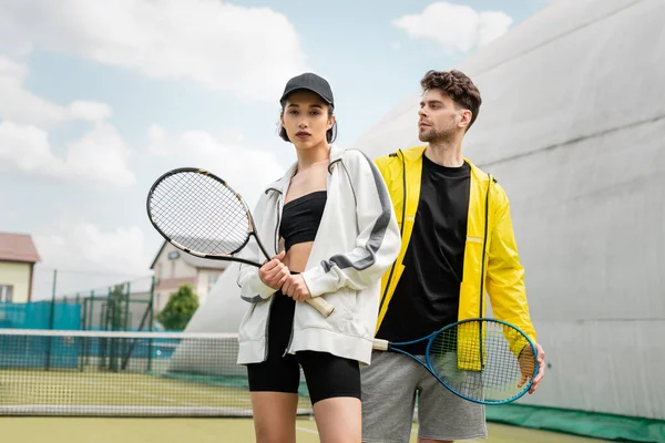 fashionable sportswear, man and woman holding tennis rackets on court, sport and style