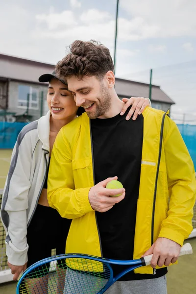 happy woman in cap and active wear hugging boyfriend holding racket and ball on court, sport