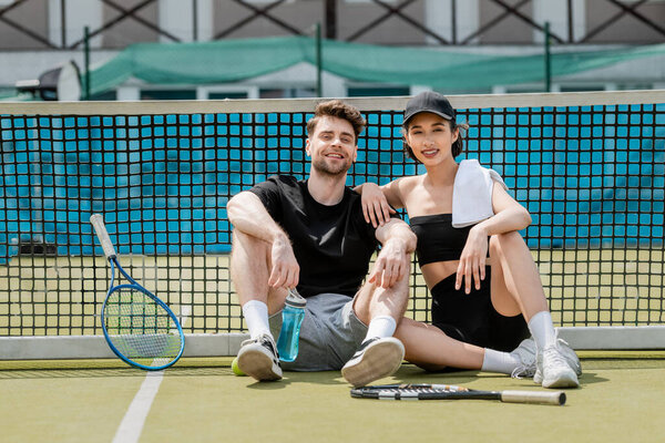 healthy lifestyle, happy man and woman in active wear resting near tennis net on court, rackets