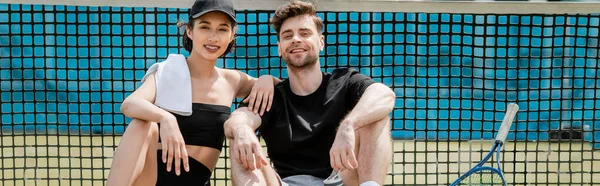 Pankart, sağlıklı yaşam tarzı, aktif durumdaki mutlu erkek ve kadın korttaki tenis ağının yanında dinleniyor.