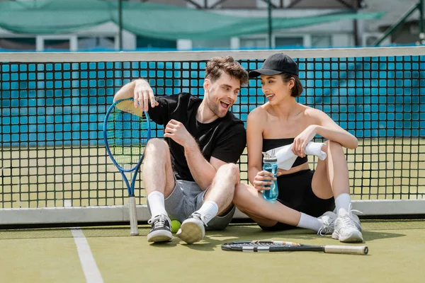 excited man and happy woman in active wear resting near tennis net on court, healthy lifestyle
