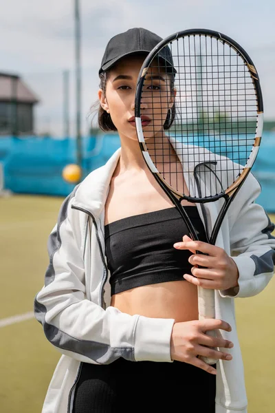 sporty woman in active wear and cap looking at camera through tennis racket on court, sport