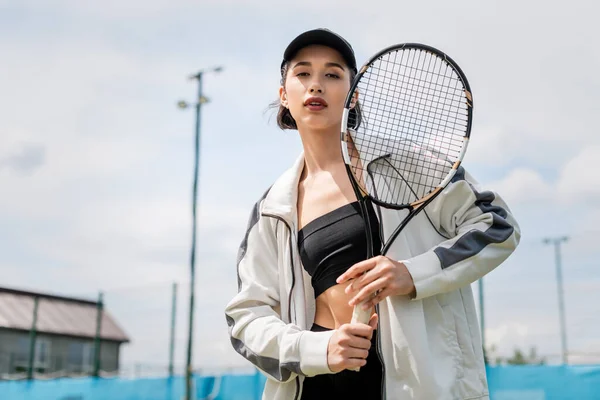 sporty woman in active wear and cap looking at camera and holding tennis racket on court, sport