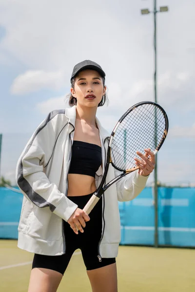 sporty woman in active wear and cap looking at camera and holding tennis racket on court, motivation
