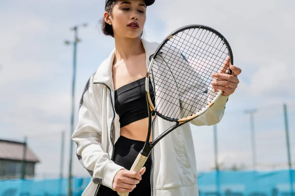 pretty woman in active wear and cap holding tennis racket on court, female tennis player, motivation
