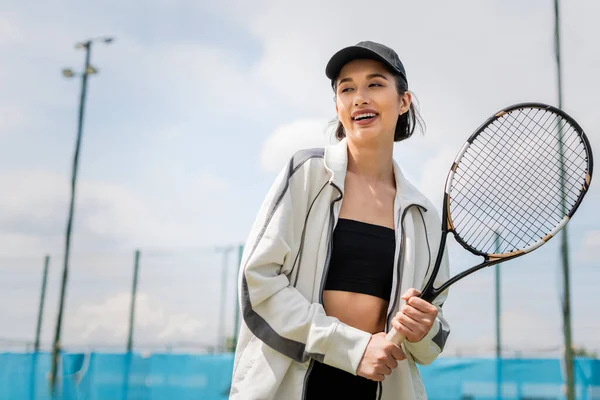 happy woman in active wear and cap holding tennis racket on court, female tennis player, motivation