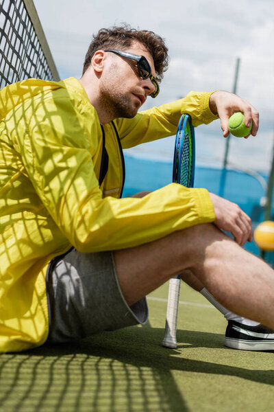 male player in sunglasses sitting near tennis net, holding ball and tennis racket, sport and style