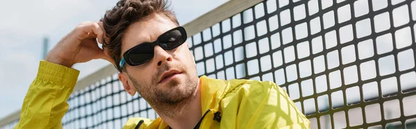 stock image banner of handsome man in sunglasses posing near tennis net, sport and style, active wear