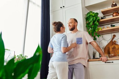 Smiling bearded man in homewear talking to girlfriend and standing together in kitchen at home clipart