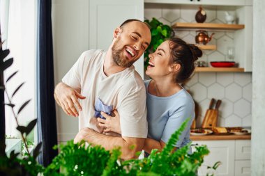 Joyful brunette woman in loungewear embracing bearded boyfriend near green plants at home clipart