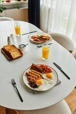 Yüksek açılı kahvaltı manzarası, kızarmış yumurta ve sosis, çatal bıçak ve portakal suyu yanında.