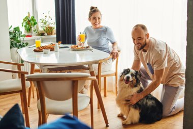 Ev kıyafetleri içinde gülümseyen bir adam sınırdaki çoban köpeğinin yanında ve evde lezzetli bir kahvaltı yapıyor.