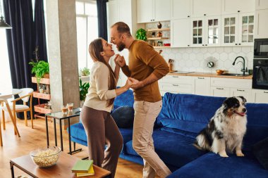 Smiling couple holding hands and dancing near border collie on couch in living room at home clipart