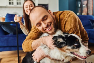 Cheerful man hugging border collie dog near blurred girlfriend with coffee in living room at home clipart