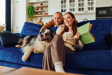 Kitaplı gülümseyen çift evde oturma odasında patlamış mısır ve kanepede Collie 'nin yanında rahatlıyor.
