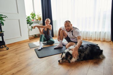 Spor giyim kuşağında gülümseyen adam, evde kız arkadaşı spor minderinde ısınırken sınır köpeğini okşuyor.