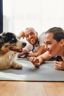 Smiling woman in sportswear lying on fitness mat near boyfriend and border collie at home clipart