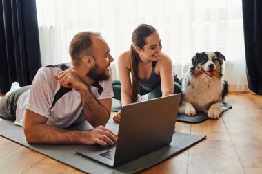 Positive couple in sportswear using laptop on fitness mats and looking at border collie at home clipart