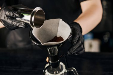 Siyah lateks eldivenli barista jigger 'dan sifon kahve makinesinin filtresine kahve döküyor.