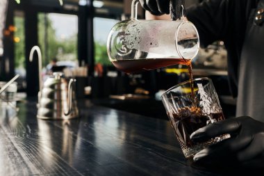 Kahve makinesini tutan baristanın kırpılmış görüntüsü ve kristal bardağa taze demlenmiş espresso dökülüyor. 