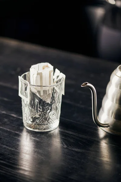 Stock image pour-over brewing method, glass with paper filter bag and metallic drip kettle on black tabletop