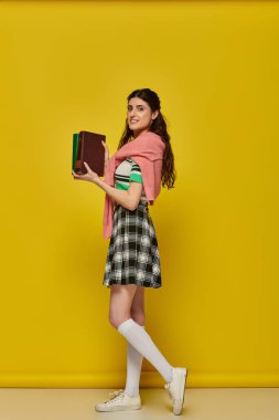 Sarı arka planda kitapları olan neşeli öğrenci, etekli genç kadın, üniversite kıyafeti
