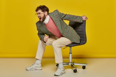 emotional and bearded man screaming and sitting on office chair, yellow backdrop, angry student
