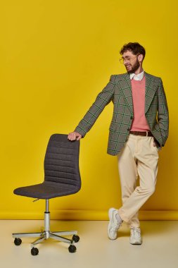 Pozitif adam, ofis sandalyesinin yanında el ele duran, sarı arka plan, gözlüklü öğrenci.