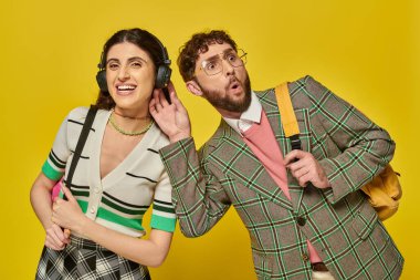 Kablosuz kulaklıklı neşeli kadın sakallı adamın yanında müzik dinliyor, öğrenciler sırt çantası tutuyor.