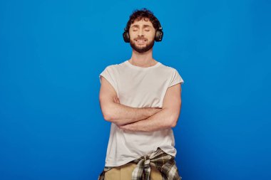 Kablosuz kulaklıklı pozitif adam mavi arka planda müzik dinliyor, kapalı gözler, yaz modası