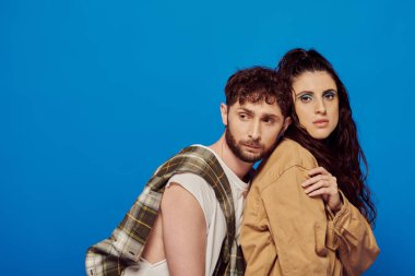 Sıradan giyinmiş şık bir çift mavi arka planda poz veriyor, sakallı bir adam kadının yanında duruyor, kalın makyajlı.