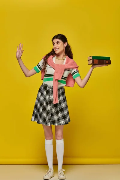 stock image happy student with books, brunette woman in checkered skirt waving hand on yellow backdrop, brunette