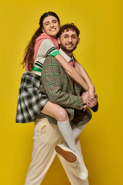 stock image couple, funny students, happy man piggybacking young woman on yellow backdrop, college outfits