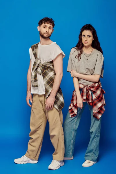 couple posing in street wear, blue backdrop, woman with bold makeup holding cap, bearded man