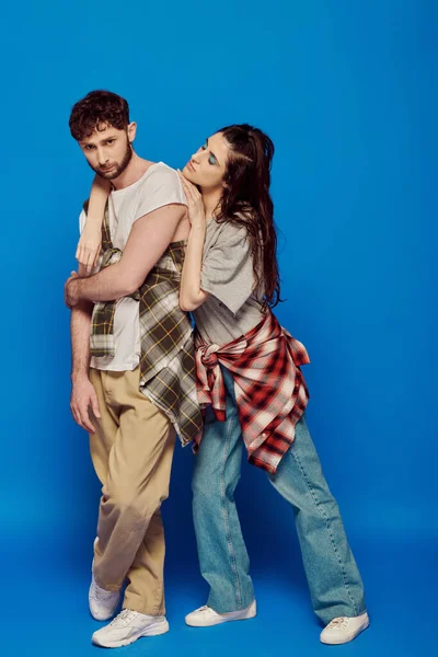 couple posing in street wear, blue backdrop, woman with bold makeup leaning on bearded man, style