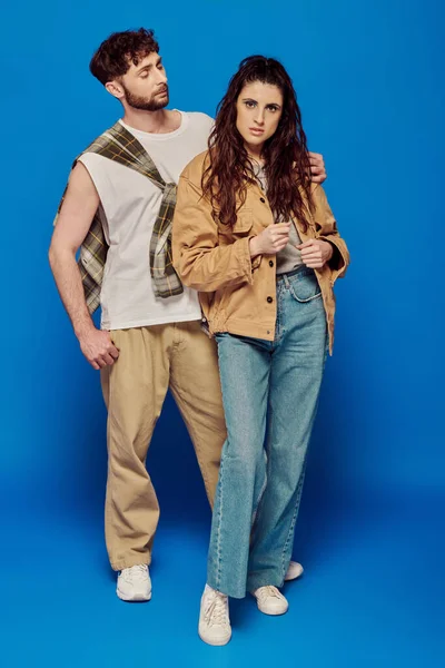 stylish couple posing in casual wear, blue backdrop, woman standing with bearded man, bold makeup