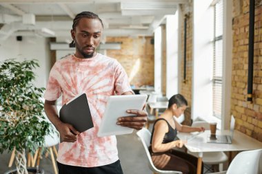 Afro-Amerikan adam tablet kullanıyor, klasörü tutuyor, kadın iş arkadaşının yanında çalışıyor, başlangıç projesi.