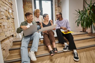 happy interracial team holding gadgets, sitting on stairs in coworking, startup, gen z modern office clipart