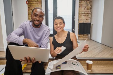 happy man and woman holding gadgets and graphs, interracial colleagues looking at camera, startup clipart