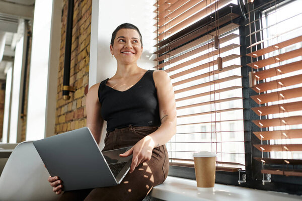 joyful tattooed woman using laptop, sitting on windowsill, coffee to go, startup project, coworking