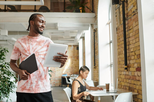 happy african american man holding tablet, folder, working near female coworker, startup project
