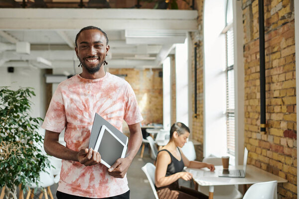 happy african american man holding tablet, folder, looking at camera near female coworker, startup