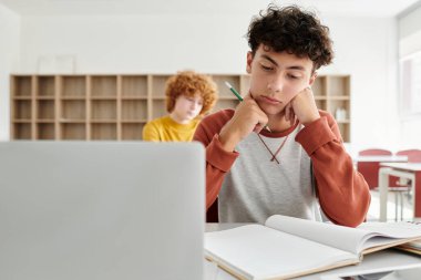 Düşünceli ergen okul çocuğu okulda dizüstü bilgisayarın ve dijital tabletin yanında deftere bakıyor.