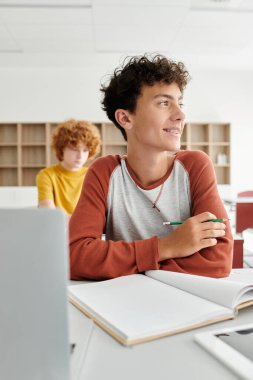 Smiling teenage schoolboy holding pencil near notebook and devices during lesson in school clipart