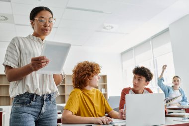 Sınıftaki öğrencilerin yakınındaki derste elinde dijital tablet tutan Afrikalı Amerikalı öğretmen.