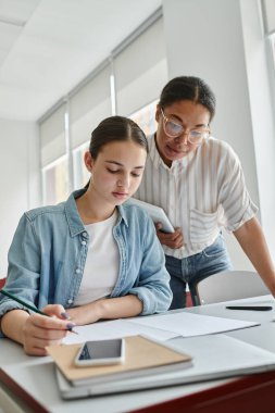 African american teacher holding digital tablet while standing near schoolgirl in classroom clipart