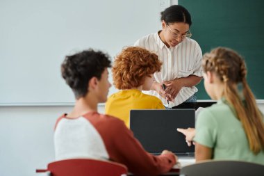 Sınıfta derste öğrencilerin yanında laptopla dikilen Afrikalı Amerikalı öğretmen.